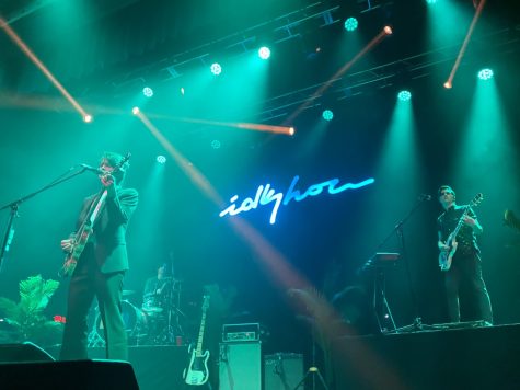 Two men on stage with idkHOW neon sign in background